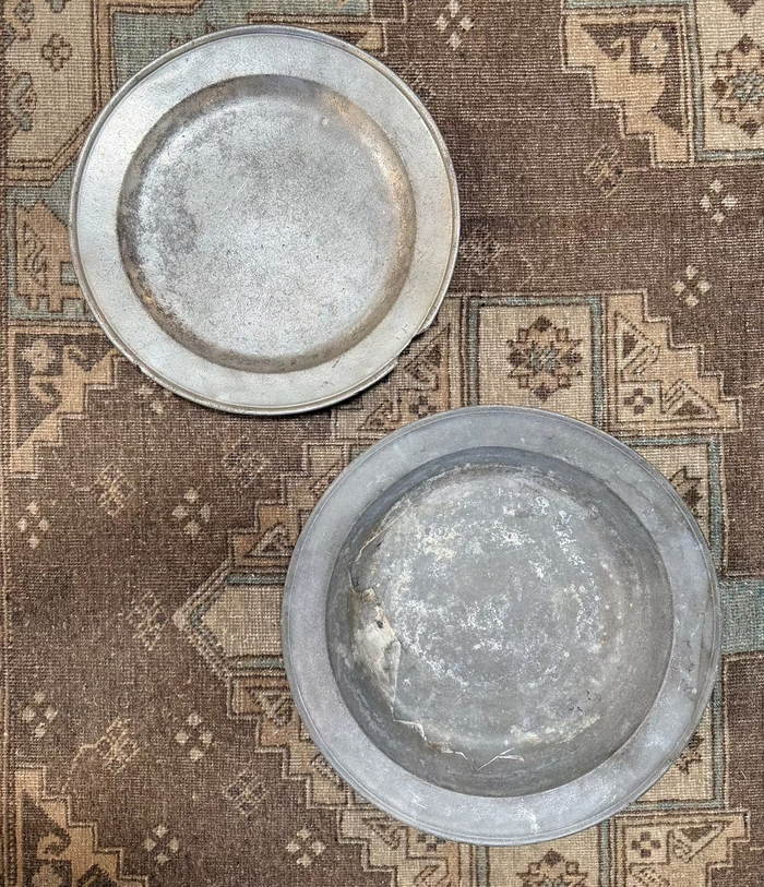 Two antique pewter platters on patterned fabric, shallow round dishes