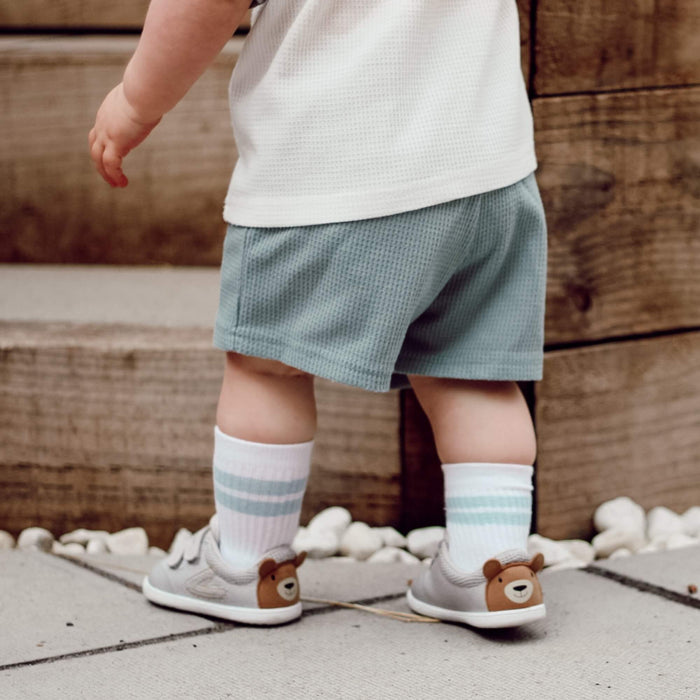 Kid's grey barefoot trainers with a brown bear face motif on the heel, pictured outdoors.