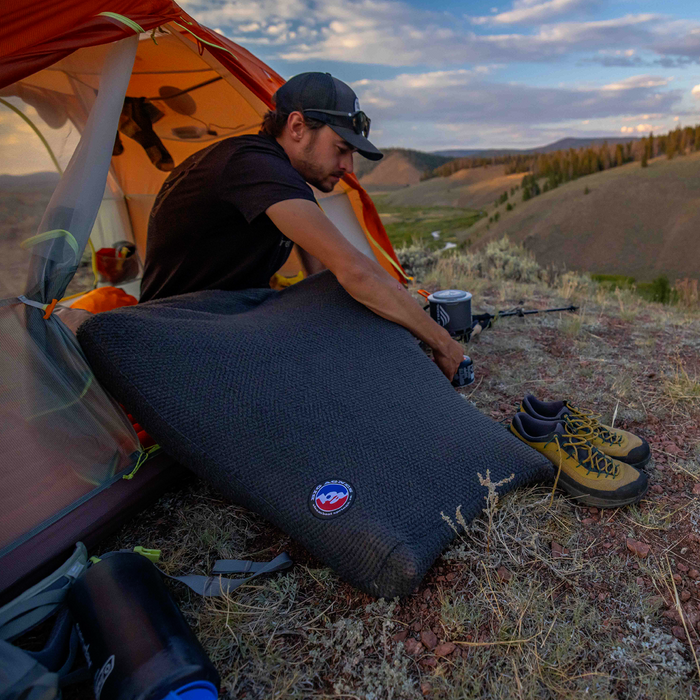 A man sits by an orange tent in hilly terrain, preparing a Big Agnes Returns Alpha Direct Fleece Sleeping Bag Liner. Hiking shoes and water bottles rest on dry grass nearby, with scenic mountains in the background.