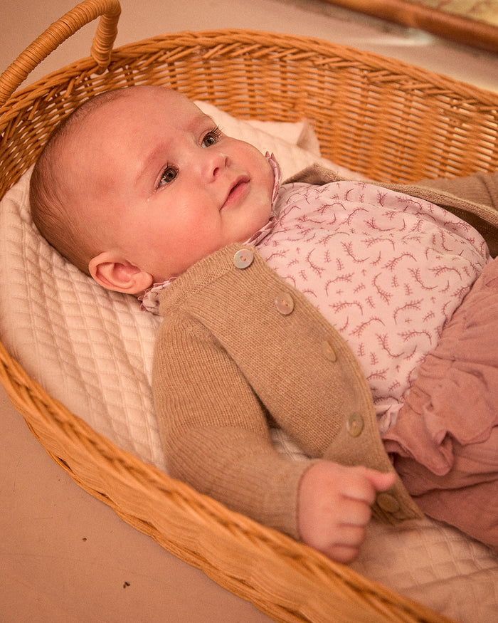 Baby in a wicker basket wearing a brown outfit and pink patterned blanket.