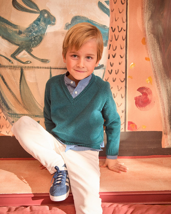 Young boy in a teal sweater and white pants sitting on a colorful rug.
