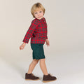 Child wearing a red plaid shirt, green shorts, and brown shoes on a plain background