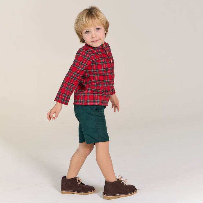 Child wearing a red plaid shirt, green shorts, and brown shoes on a plain background