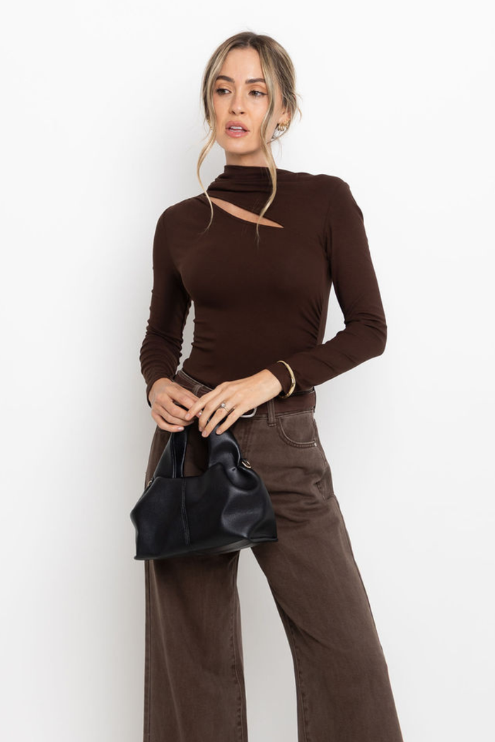 Woman wearing a brown long-sleeve top and pants holding a black handbag against a white background
