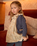 Young girl wearing a patterned dress and blue cardigan standing in front of a red couch with pillows.
