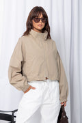 Woman wearing a stone funnel neck bomber jacket over white pants, styled with a brown suede bag, on a white background.