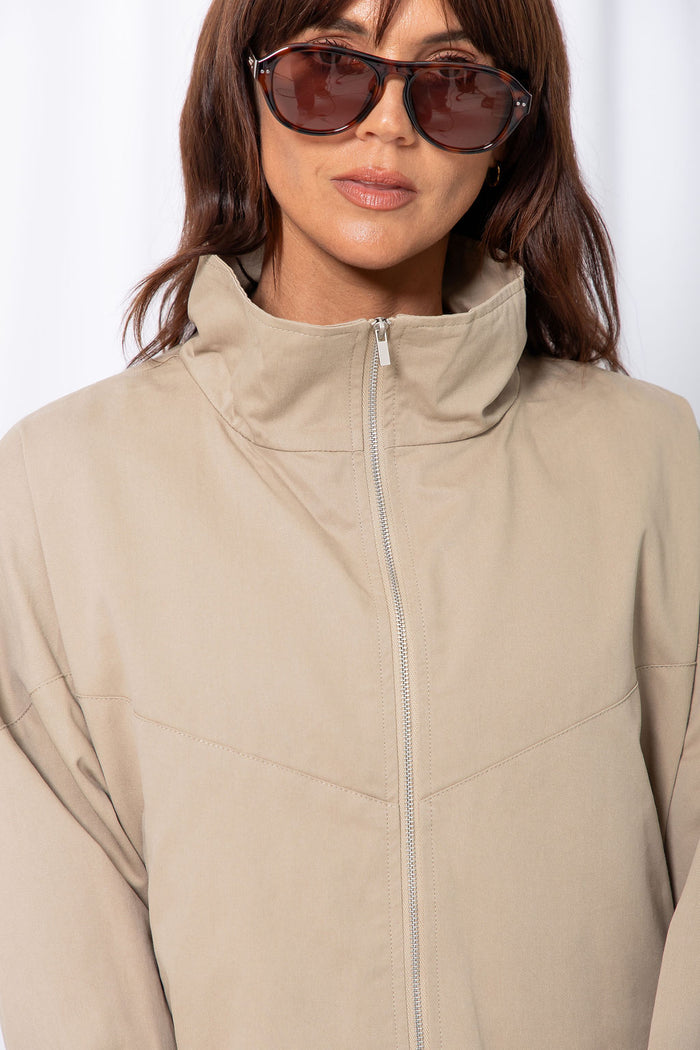 Woman wearing a stone funnel neck bomber jacket over white pants, styled with a brown suede bag, on a white background.