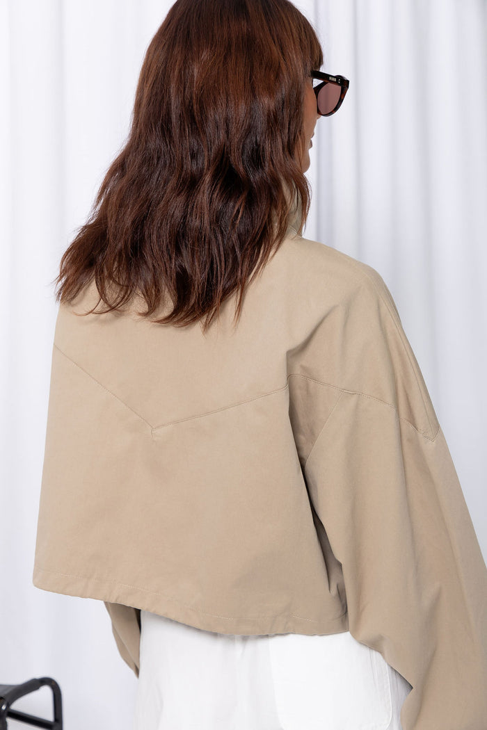 Woman wearing a stone funnel neck bomber jacket over white pants, styled with a brown suede bag, on a white background.