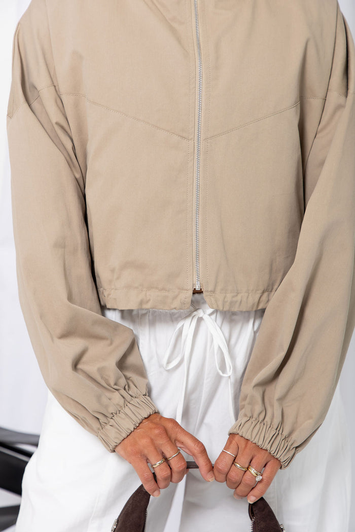 Woman wearing a stone funnel neck bomber jacket over white pants, styled with a brown suede bag, on a white background.