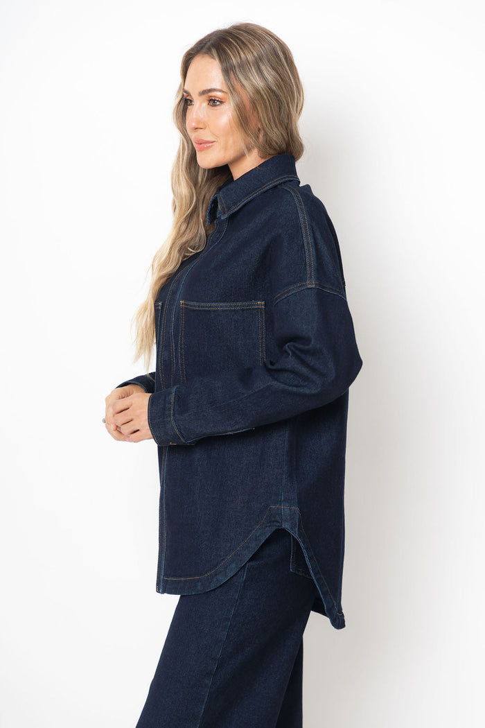 A woman wearing an oversized dark indigo denim shirt jacket with a pointed collar and front chest pocket, shown in profile against a plain white studio background.