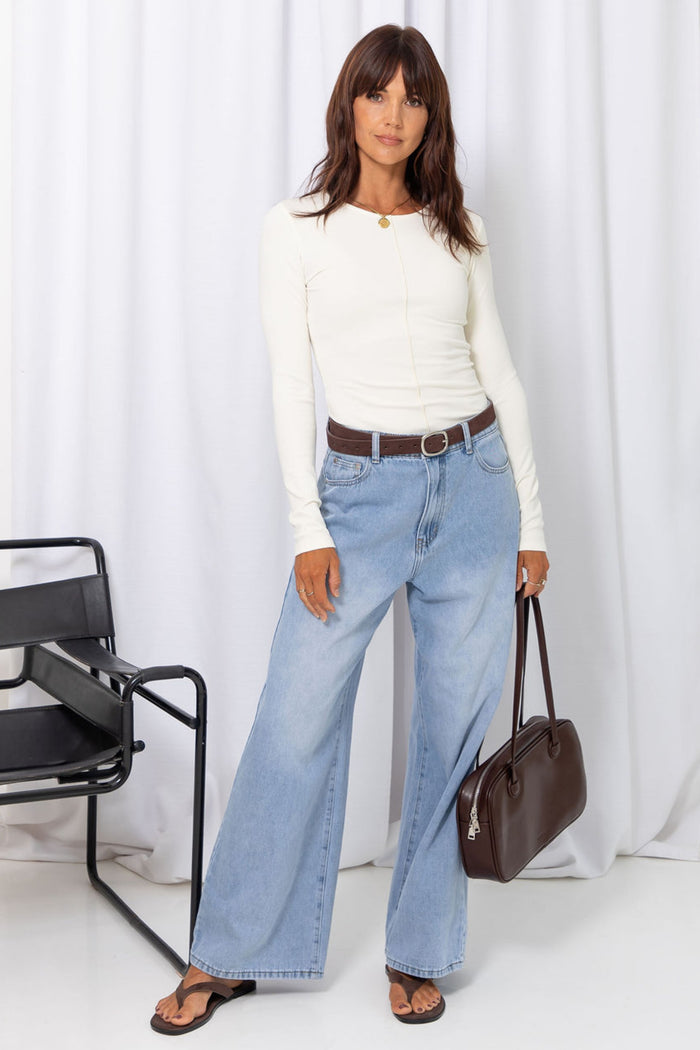 Woman wearing a white long-sleeve top with light-wash wide-leg jeans and a brown belt, carrying a brown handbag, on a white background.