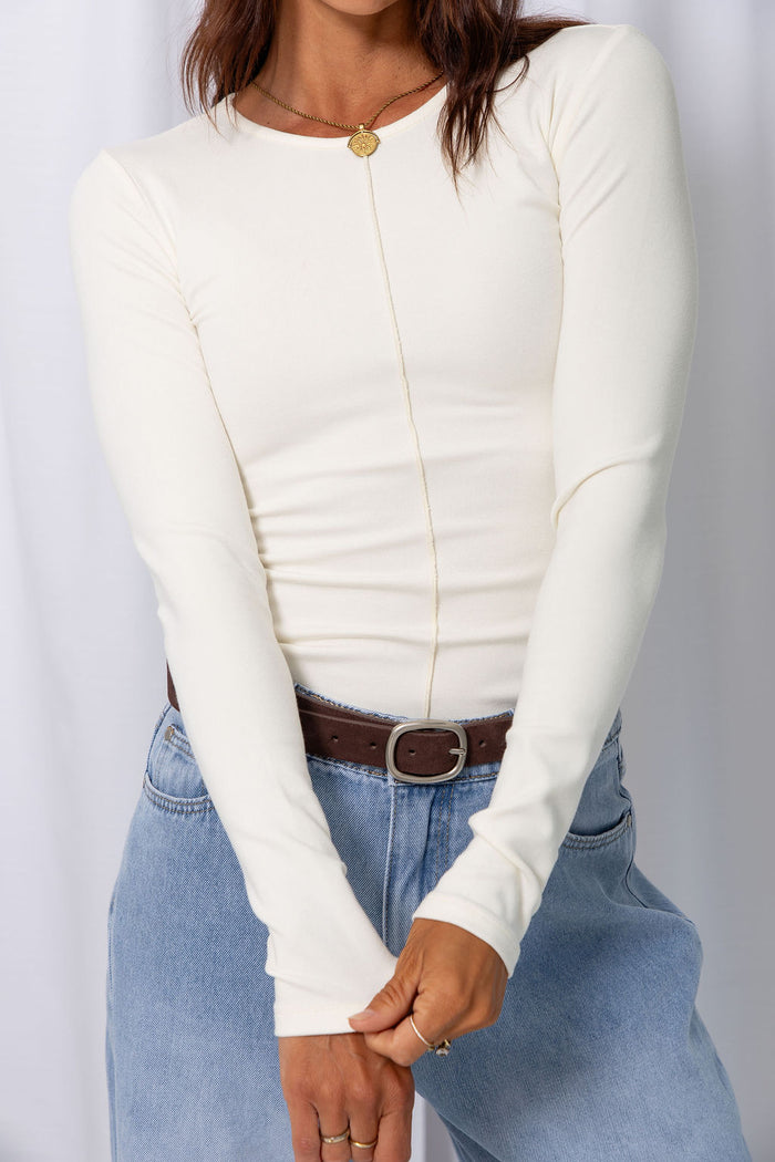 Woman wearing a white long-sleeve top with light-wash wide-leg jeans and a brown belt, carrying a brown handbag, on a white background.