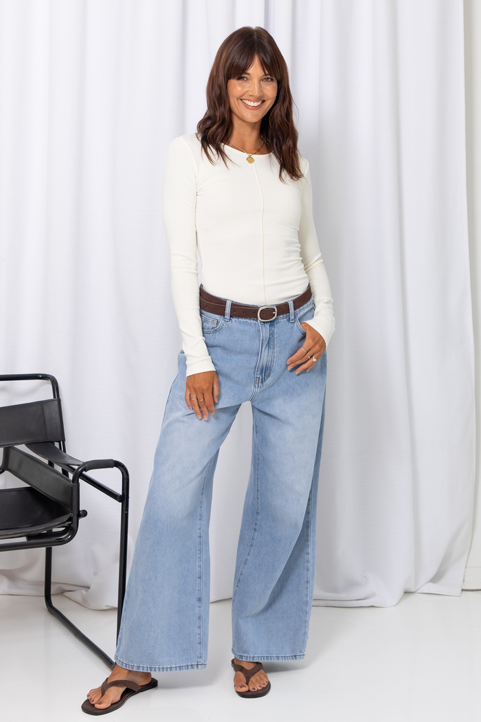 Woman wearing a white long-sleeve top with light-wash wide-leg jeans and a brown belt, carrying a brown handbag, on a white background.