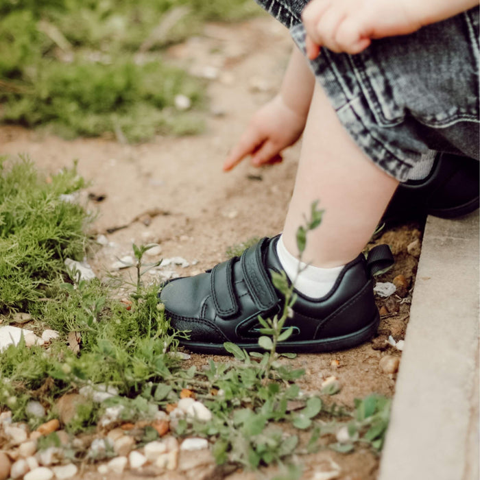 Toddler wearing black barefoot trainers sitting in park close up of shoes.