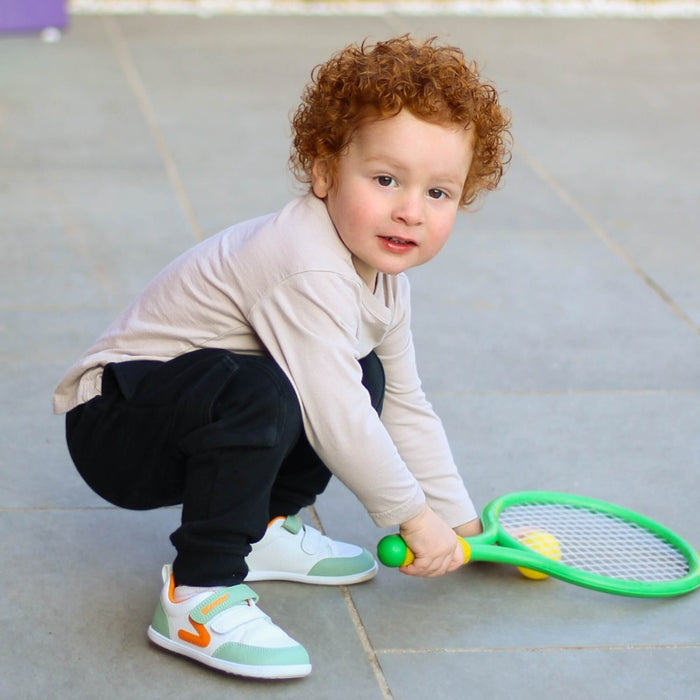 Kid wearing white and olive green barefoot trainers playing with a toy racket on an outdoor patio.