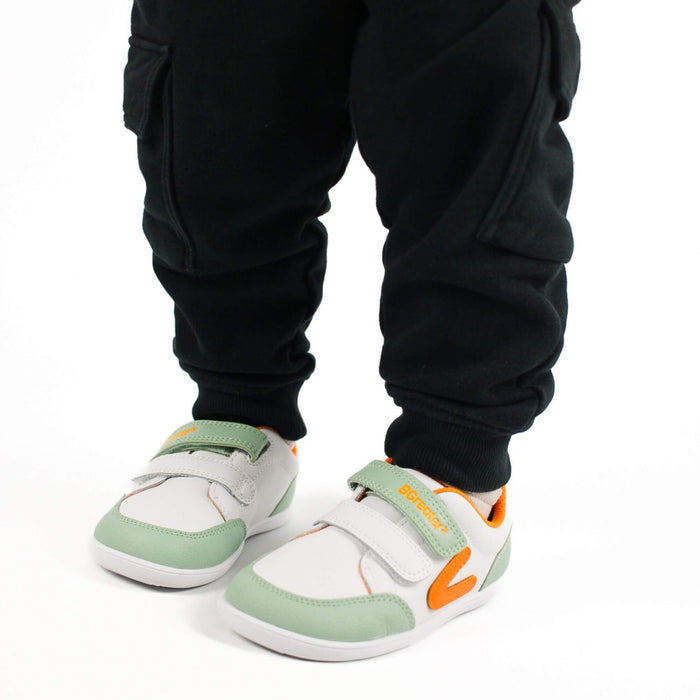 Close-up of a kid wearing white and olive green barefoot trainers with hook-and-loop straps on a white background.