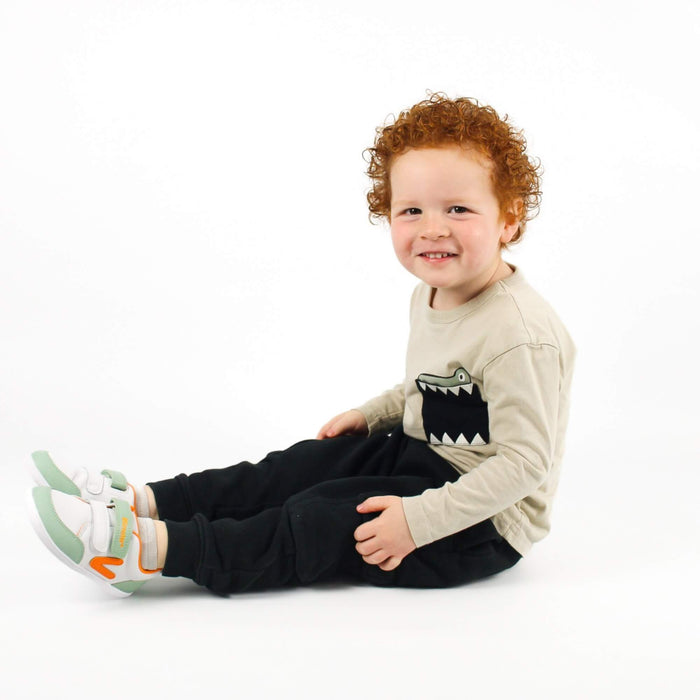Smiling kid sitting on a white studio background wearing white and olive green barefoot trainers.