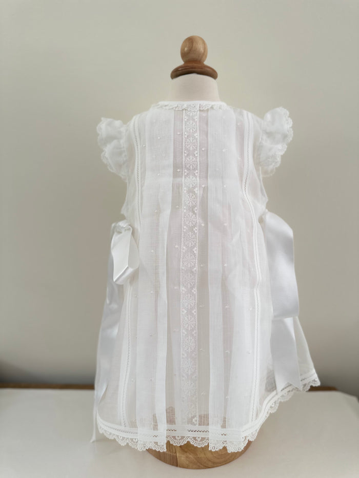 A white dress for a girl with satin bows on the sides and lace detailing, displayed on a mannequin. Made by Spanish brand, Creaciones Charo.