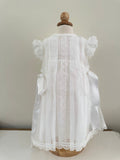 A white dress for a girl with satin bows on the sides and lace detailing, displayed on a mannequin. Made by Spanish brand, Creaciones Charo.