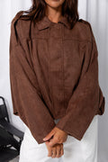 Woman wearing a brown suede-look jacket over a white top and white pants, standing against a white curtain background.