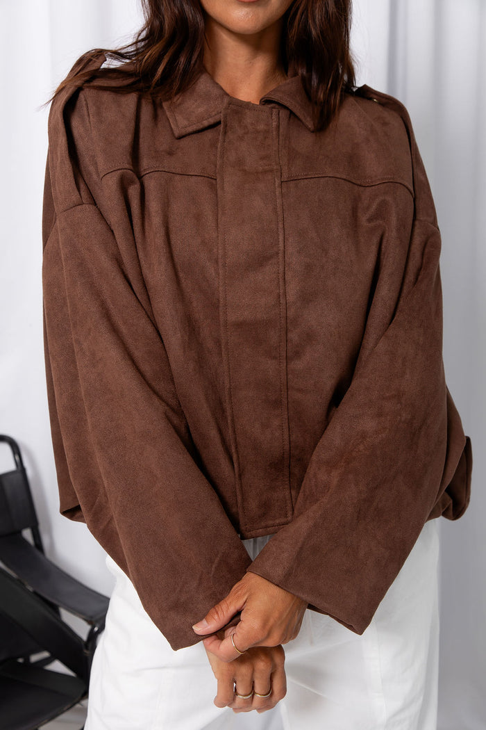 Woman wearing a brown suede-look jacket over a white top and white pants, standing against a white curtain background.