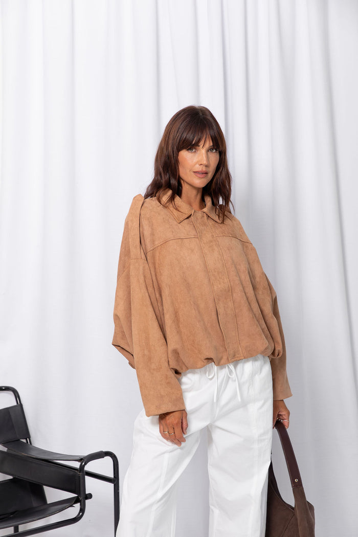 Woman wearing a sand suede-look jacket over a white top and white pants, standing against a white curtain background.