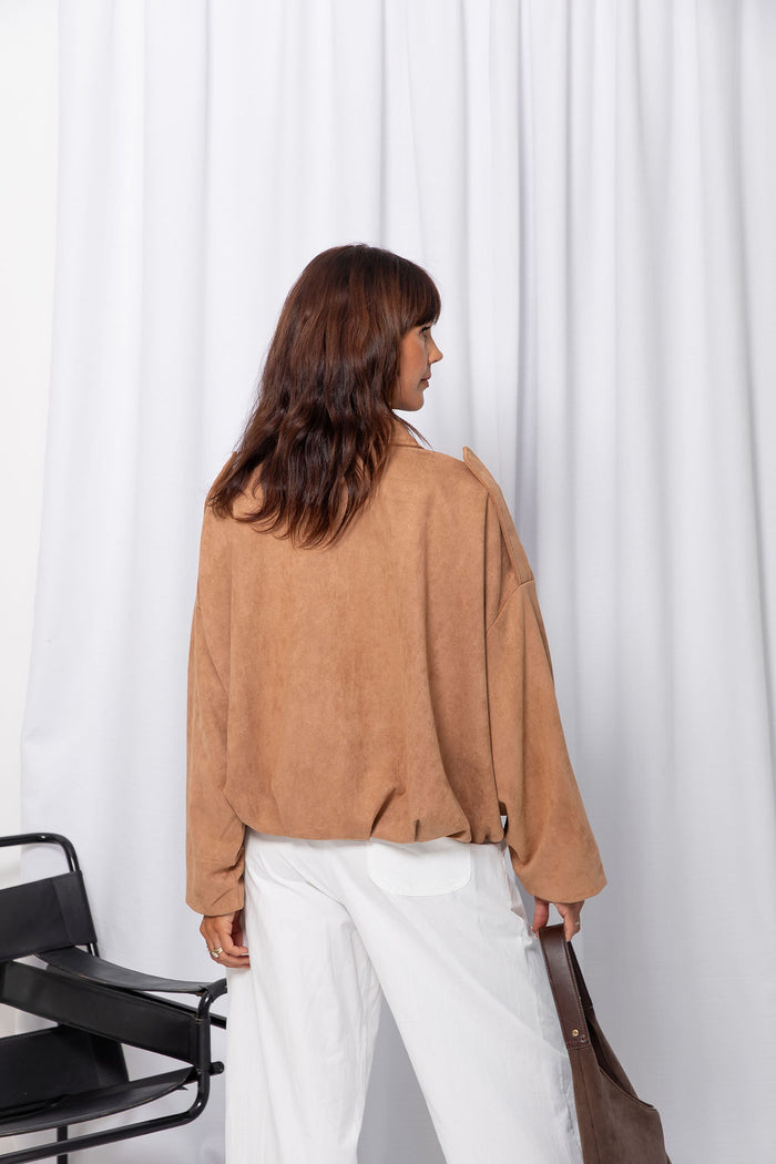 Woman wearing a sand suede-look jacket over a white top and white pants, standing against a white curtain background.