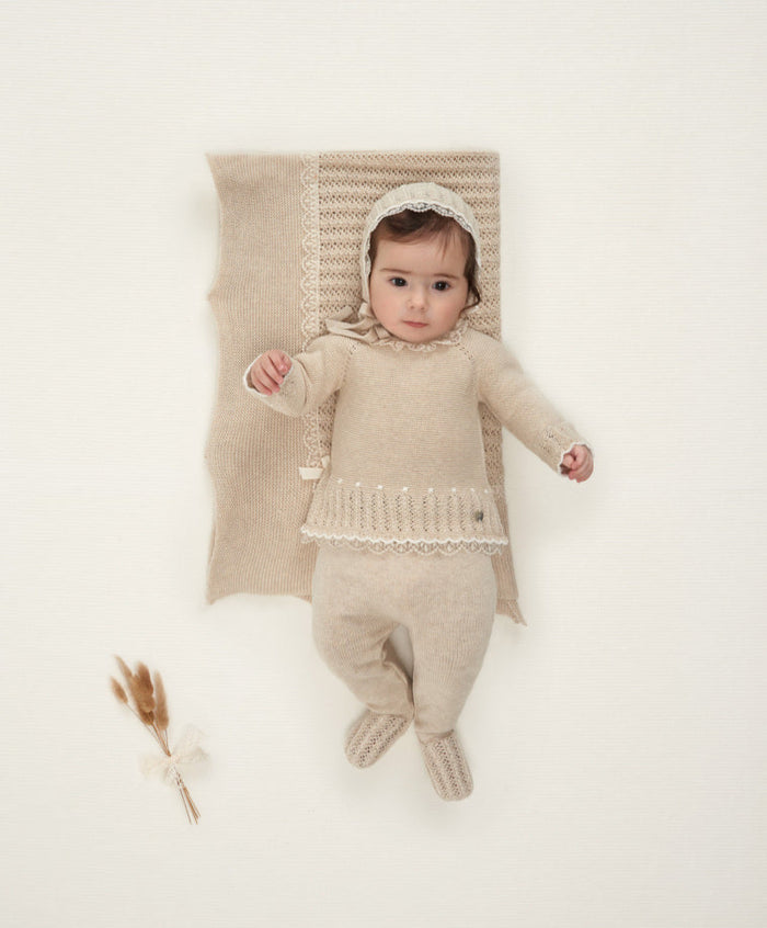Baby in a beige knitted outfit lying on a white blanket with a textured pillow.