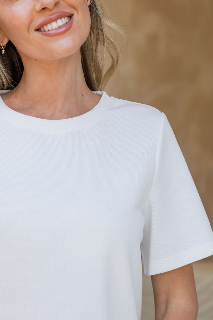 A close-up of a woman wearing a plain white short-sleeve crewneck t-shirt, cropped from the mouth down against a warm beige textured background.