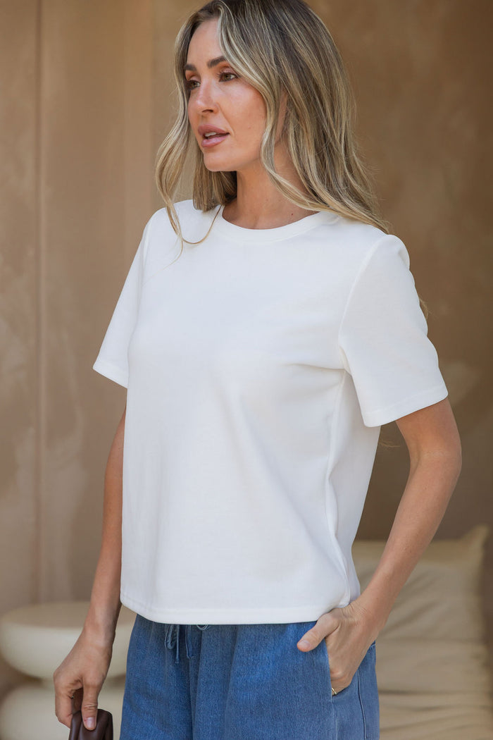 A woman wearing a white short-sleeve crewneck t-shirt and blue jeans, standing against a neutral beige background.