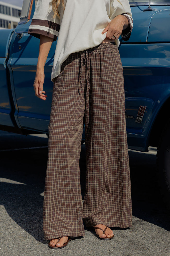 A person wearing brown and beige plaid wide-leg drawstring pants, standing outdoors next to a blue pickup truck.
