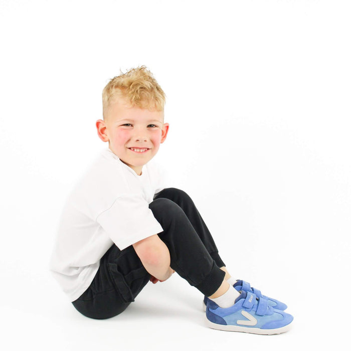 Smiling kid sitting on a white studio background wearing blue leather barefoot trainers.