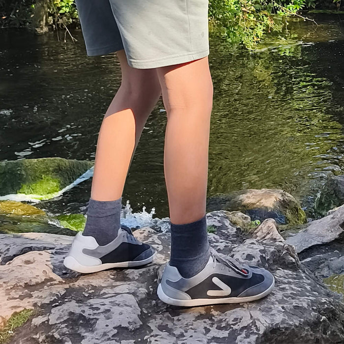 Kid's grey and black barefoot trainers standing on outdoor rocks by water.