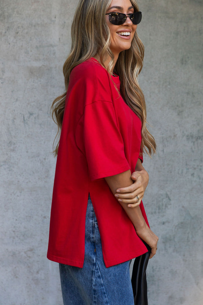 A woman wearing an oversized bright red short-sleeve top and blue jeans, posing against a textured gray concrete wall.