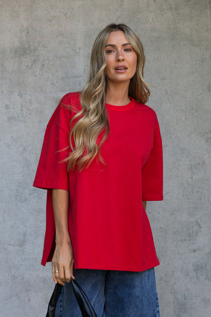 A woman with blonde wavy hair wearing a bright red oversized short-sleeve top and blue jeans, standing against a textured gray concrete wall.