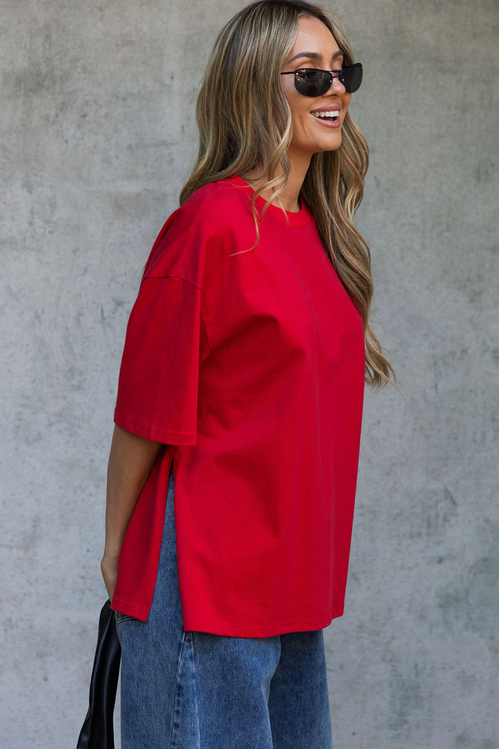 A woman wearing an oversized bright red short-sleeve top and blue jeans, standing against a textured gray concrete wall.