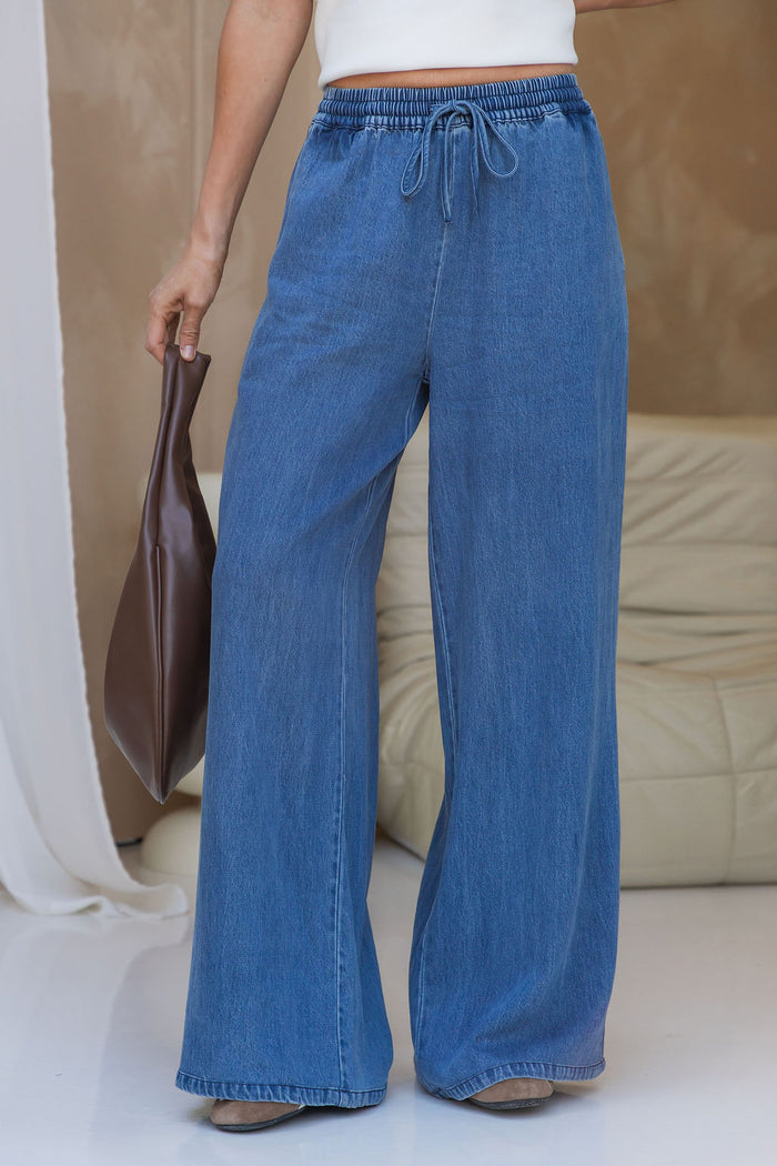 A person wearing wide-leg blue denim drawstring pants, holding a brown leather bag, standing in a brightly lit indoor setting with a beige sofa in the background.