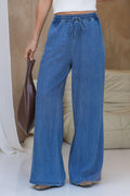 A person wearing wide-leg blue denim drawstring pants, holding a brown leather bag, standing in a brightly lit indoor setting with a beige sofa in the background.
