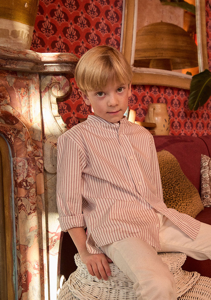 Young boy sitting on a wicker stool in a room with patterned wallpaper and a red couch.