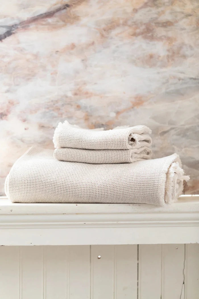 Light ecru waffle hand towel set with fringed edges, neatly stacked on a shelf.