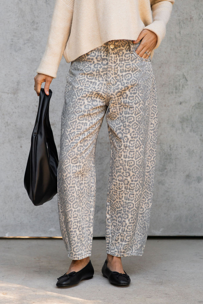 A person wearing loose, wide-leg trousers with a beige and gray animal print pattern, paired with a cream sweater and black flats, holding a black handbag against a gray concrete wall.